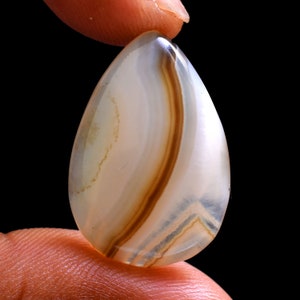 May include: A white agate cabochon with brown and gray banding. The cabochon is teardrop shaped and has a smooth, polished surface.