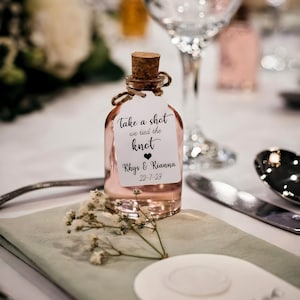 May include: A small glass bottle with a cork stopper filled with a pink liquid. A white tag is attached to the bottle with the text "Take a shot we tied the knot Rhys & Rianna 22-7-23" and a small heart symbol.
