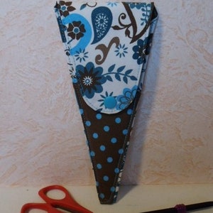 May include: A fabric scissor case with a blue and white floral pattern and a brown polka dot base. A pair of red-handled scissors and two black knitting needles are in the foreground.