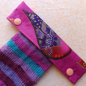 Op de afbeelding: Een close-up van een handgemaakt, levendig paars en roze stoffen item met een knoopsluiting, naast een gestreepte gebreide sok in tinten paars, blauw en roze. De stof heeft een decoratief ontwerp met paarse en gouden accenten.