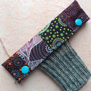 Könnte beinhalten: Ein handgefertigter Handgelenkwärmer mit einem gemusterten Stoffpaneel und einer gestrickten Manschette. Das Stoffpaneel hat ein Patchwork-Design mit orangefarbenen, lila und grünen Mustern, das mit zwei türkisfarbenen Knöpfen befestigt ist. Die gestrickte Manschette ist grau-grün.