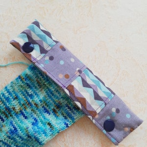 Könnte beinhalten: Ein gestrickter Socken in verschiedenen Blau- und Grüntönen, der an einer Stoffmanschette befestigt ist. Die Manschette zeigt ein geometrisches Zickzackmuster in Lila, Blau und Weiß, das sich mit einem lila Stoffeinsatz mit bunten Punkten abwechselt.