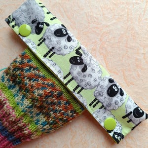 Könnte beinhalten: Ein grüner und weißer Stoff-Maschenmarkierer mit einem Schaf-Muster und lindgrünen Knöpfen. Der Maschenmarkierer liegt auf einer bunten gestrickten Socke.