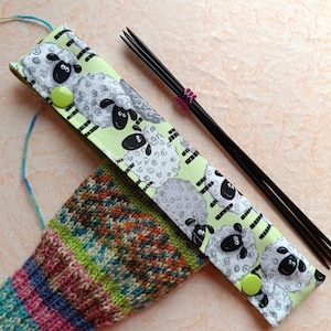 Könnte beinhalten: Eine grüne Nadeltasche aus Stoff mit einem Schaf-Muster und schwarzen Knöpfen. In der Tasche befinden sich schwarze Stricknadeln mit einem lilafarbenen Maschenmarkierer.