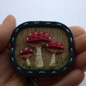 The mushroom brooch is shown with somebody holding it. Made from felt and wood with handstitched detail. There are three different sized mushrooms with red tops and surrounded by blades of grass.