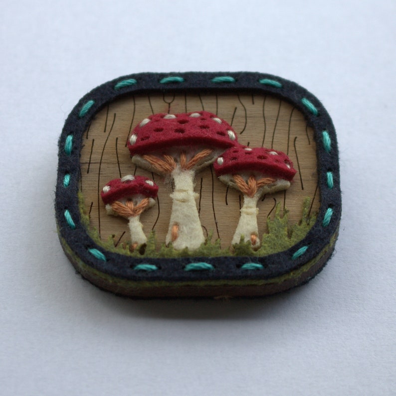 Close up view of the mushroom brooch made from walnut veneer wood with felt pieces hand stitched on. There are three red mushrooms, green grass in the foreground and the woodland scene is framed with a dark blue felt surround.
