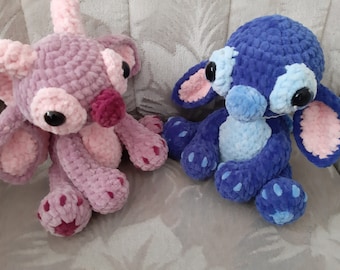 May include: Two crocheted plush toys, one pink and one blue, with large eyes and stitched details. The toys are sitting on a light gray surface.