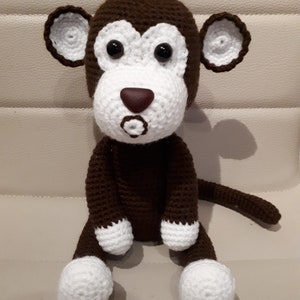 Può includere: Un giocattolo di scimmia all'uncinetto marrone con accenti bianchi. La scimmia ha un viso bianco con occhi marroni e un naso rosso. La scimmia è seduta con le gambe incrociate e la coda arrotolata dietro di lei.