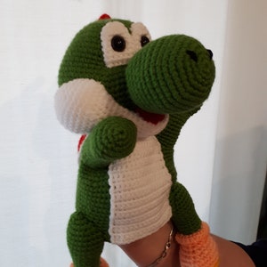 Könnte beinhalten: Grünes Yoshi Plüschtier aus Häkelarbeit mit weißen und orangefarbenen Akzenten. Das Spielzeug hat einen roten, spitz zulaufenden Kamm auf dem Kopf und schwarze Augen.