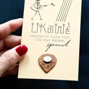 Puede incluir: Un primer plano de una púa de guitarra de madera marrón con un centro blanco y el texto "Little Axe" grabado en ella. La púa está en una tarjeta con el texto "Little Axe Handcrafted Guitar Picks For Your Maximal Speed".