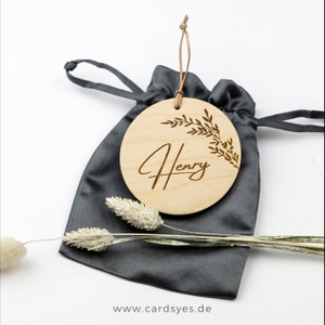 Pu&ograve; includere: Un ornamento rotondo in legno con un design a ramo frondoso e il nome "Henry" inciso in scrittura corsiva. L'ornamento &egrave; in una borsa a cordoncino grigia.