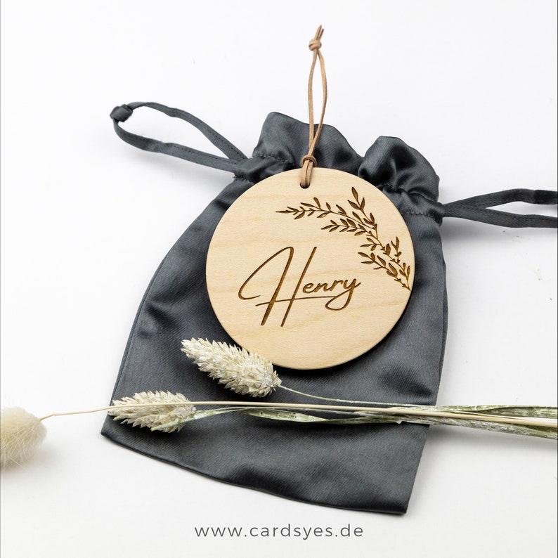 Pu&ograve; includere: Un ornamento rotondo in legno con un design a ramo frondoso e il nome "Henry" inciso in scrittura corsiva. L'ornamento &egrave; in una borsa a cordoncino grigia.