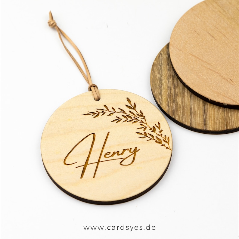 Pu&ograve; includere: Un ornamento rotondo in legno di colore marrone chiaro con un cordino in pelle. L'ornamento ha un design floreale e il nome "Henry" inciso su di esso.