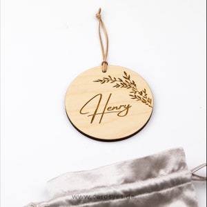 Pu&ograve; includere: Un ornamento rotondo in legno con un cordino in pelle marrone. L'ornamento &egrave; inciso con il nome "Henry" in un carattere corsivo e un disegno di foglie.
