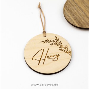 Pu&ograve; includere: Un ornamento rotondo in legno con una finitura marrone chiaro. L'ornamento presenta un delicato motivo floreale e il nome "Henry" inciso in un carattere corsivo. L'ornamento &egrave; attaccato a un cordino in pelle marrone.