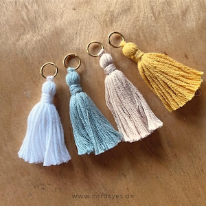 Op de afbeelding: Vier kleurrijke kwastjes sleutelhangers in wit, blauw, beige en geel, elk met een goudkleurige ring. De kwastjes zijn gemaakt van zacht garen en zijn gerangschikt op een houten oppervlak. Het websiteadres "cardsyes.de" is zichtbaar linksonder.