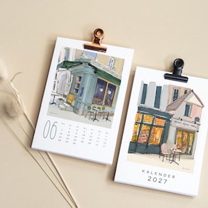 Può includere: Due pagine di calendario da parete con illustrazioni in stile acquerello di vetrine. La pagina sinistra mostra giugno con una facciata verde, mentre la destra riporta il testo KALENDER 2027 con un'illustrazione. Entrambe fissate con clip metalliche.