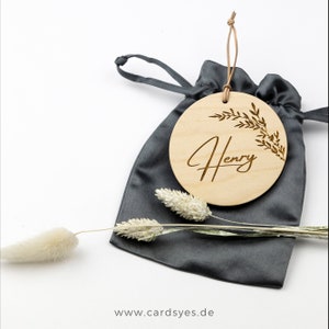 Pu&ograve; includere: Un ornamento rotondo in legno con il nome "Henry" inciso in corsivo. L'ornamento &egrave; decorato con un motivo di ramo frondoso e viene fornito in una borsa di cordoncino grigia.