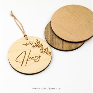 Pu&ograve; includere: Un ornamento rotondo in legno con il nome "Henry" inciso in corsivo. L'ornamento &egrave; decorato con un motivo di ramo frondoso e ha un cordino in pelle marrone per appenderlo.