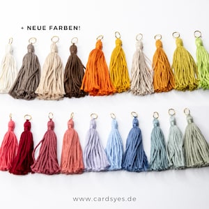 Könnte beinhalten: Eine Sammlung von 20 bunten Quasten-Schlüsselanhängern mit goldenen Ringen. Die Quasten sind aus Garn in verschiedenen Farben gefertigt, darunter Rot, Orange, Gelb, Grün, Blau, Lila, Rosa und Braun. Der Text '+ NEUE FARBEN!' ist oben im Bild sichtbar.