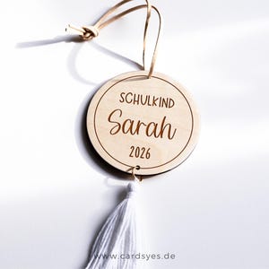 Ein runder Holzanhänger mit einer weißen Quaste. Der Anhänger ist mit dem Text "Schulkind 2026“ graviert.