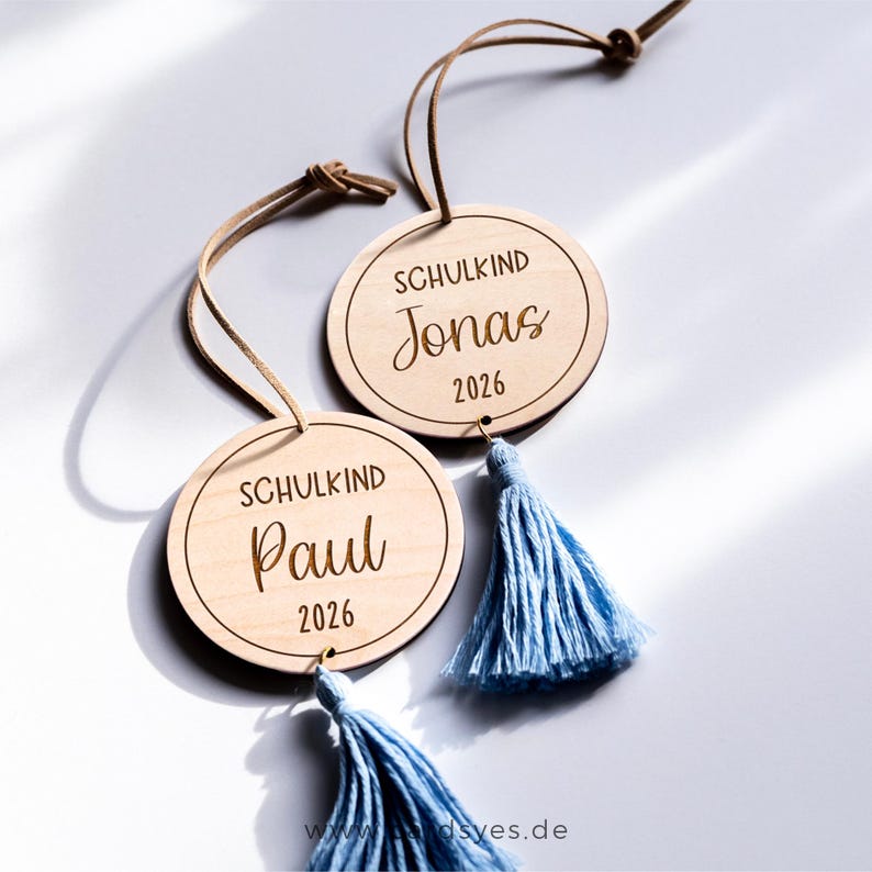 Zwei runde Holzornamente mit blauen Quasten. Die Holzanhänger sind mit dem Text "Schulkind" und den Namen "Jonas" und "Paul" sowie dem Jahr "2026" graviert.