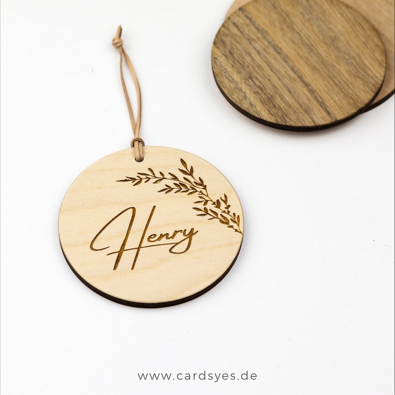 Pu&ograve; includere: Un ornamento rotondo in legno con una finitura marrone chiaro. L'ornamento ha un cordino in pelle per appenderlo e presenta un motivo floreale e il nome "Henry" inciso in un carattere corsivo.