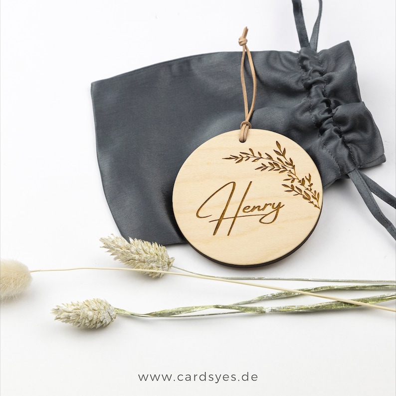 Pu&ograve; includere: Un ornamento rotondo in legno con il nome "Henry" inciso in corsivo. L'ornamento &egrave; decorato con un motivo a foglie e viene fornito con un sacchetto di cordoncino grigio.