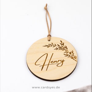 Pu&ograve; includere: Un ornamento rotondo in legno con una finitura in legno naturale. L'ornamento presenta un delicato disegno floreale e il nome "Henry" inciso in un carattere corsivo. L'ornamento &egrave; attaccato a un cordoncino in pelle marrone.