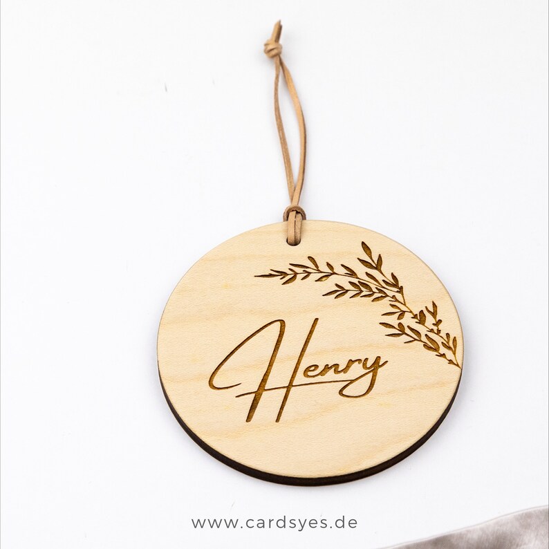 Pu&ograve; includere: Un ornamento rotondo in legno con una finitura in legno naturale. L'ornamento presenta un delicato disegno floreale e il nome "Henry" inciso in un carattere corsivo. L'ornamento &egrave; attaccato a un cordoncino in pelle marrone.