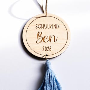 Holzanhänger mit Quasten. Der Anhänger ist mit dem Text "Schulkind 2026“ graviert.