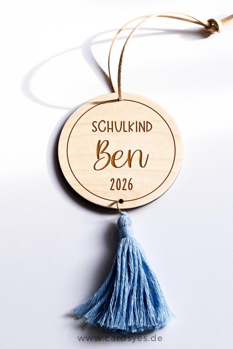 Holzanhänger mit Quasten. Der Anhänger ist mit dem Text "Schulkind 2026“ graviert.
