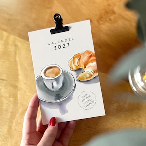 Può includere: Una scheda calendario 2027 con un'illustrazione ad acquerello di una tazza di caffè su un piattino e un cornetto dorato. Il testo recita KALENDER 2027 e JOY IN THE LITTLE THINGS. Una clip in metallo nero è fissata sulla parte superiore della carta bianca.