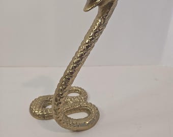 Könnte beinhalten: Eine goldfarbene Kobra-Statue mit offenem Maul, bereit zum Zustoßen. Der Körper der Schlange ist gewunden und ihre Schuppen sind detailliert. Die Statue ist aus Metall und hat eine glänzende Oberfläche. Die Kobra ist ein dekorativer Gegenstand.