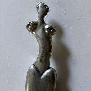 May include: A silver-toned metal brooch shaped like a stylized human figure. The piece features an elongated torso, a small head with subtle facial features, and circular chest details. The surface has a smooth, polished finish with a metallic sheen.