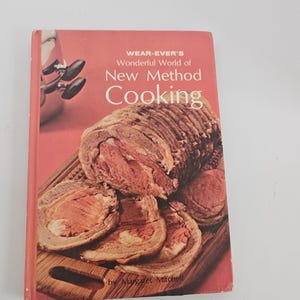May include: A vintage cookbook titled "Wear-Ever's Wonderful World of New Method Cooking" by Margaret Mitchell. The cover is salmon-colored with a photo of sliced roast beef on a wooden cutting board. The book's title is in white lettering.