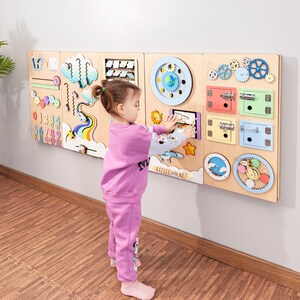 Könnte beinhalten: Ein Kind interagiert mit einer hölzernen sensorischen Tafel, die an einer Wand befestigt ist. Die Tafel verfügt über verschiedene interaktive Elemente, darunter Zahnräder, Riegel und Formen. Die Tafel besteht aus hellem Holz und hat farbige Akzente.