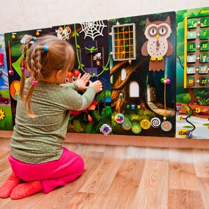 Pode incluir: Uma crian&ccedil;a brinca com um painel de atividades de madeira colorido que inclui um jogo de mem&oacute;ria, um rel&oacute;gio, engrenagens e um labirinto. O painel est&aacute; montado em uma parede e tem uma combina&ccedil;&atilde;o de cores verde, marrom e amarelo.