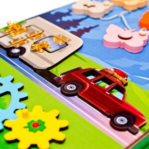 Pode incluir: Um brinquedo de madeira com um carro vermelho, um autocaravana e engrenagens. O carro tem um porta-bagagens no telhado com bagagem no topo. A autocaravana tem uma porta com uma trava. As engrenagens s&atilde;o azuis, amarelas e vermelhas. O fundo &eacute; verde e azul.