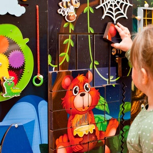 Pode incluir: Uma crian&ccedil;a brinca com um painel de atividades de madeira colorido que apresenta uma variedade de elementos interativos, incluindo um sistema de engrenagens, uma lagarta, uma barraca, uma teia de aranha e um quebra-cabe&ccedil;a com um urso segurando um pote de mel. O painel est&aacute; montado em um fundo preto.