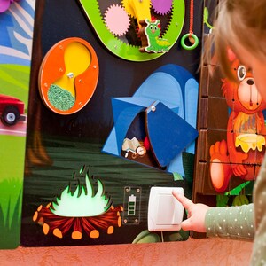 Pode incluir: Uma crian&ccedil;a interage com um painel de atividades de madeira colorido que apresenta um carro, uma barraca, uma fogueira, engrenagens e um interruptor de luz. O painel &eacute; projetado para que as crian&ccedil;as aprendam e brinquem.