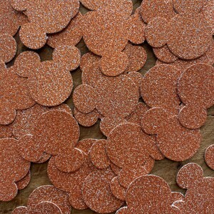 Puede incluir: Una colección de confeti con forma de cabeza de Mickey Mouse, de color cobre brillante, esparcido sobre una superficie de madera. El confeti tiene un aspecto texturizado y brillante, ideal para decoraciones de fiestas o proyectos de manualidades.