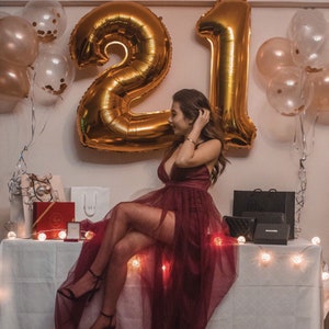 May include: A woman in a burgundy dress with a sheer overlay sits on a table with a white tablecloth.  She is surrounded by gold balloons that spell out "21".