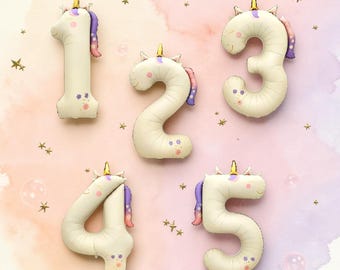 May include: A set of five cream-coloured foil balloons shaped like the numbers one through five. Each balloon features a unicorn head with a gold horn, a purple and pink mane, and small floral accents in orange, purple, and blue colours.