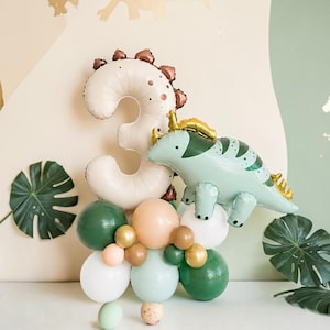 May include: A birthday decoration with a number 3 balloon, a dinosaur balloon, and a cluster of balloons in various colours. Green monstera leaves and gold dinosaur silhouettes complete the party theme.