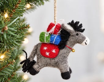 May include: A felt donkey ornament in grey with black hooves and mane. It carries a red saddle with white snowflake embroidery and three small gift boxes in red, green, and blue on its back. A small gold bell hangs from a red collar.