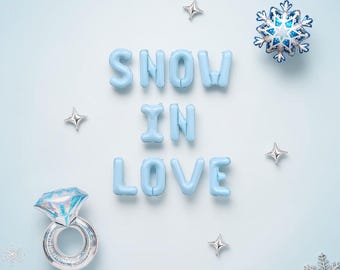 May include: Light blue foil balloons spelling the phrase SNOW IN LOVE, accompanied by a large diamond ring foil balloon, four small silver star balloons, and two silver snowflake balloons on a light blue background.