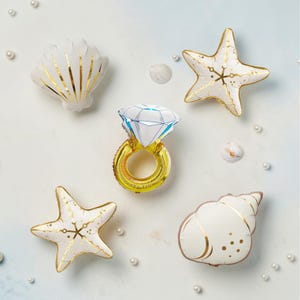 May include: A collection of five foil balloons featuring a central diamond ring balloon with a gold band and white stone, surrounded by two white and gold starfish balloons and two white and gold seashell balloons, accented by small white pearls.