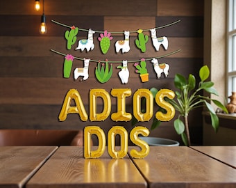 May include: A festive party decoration featuring gold foil balloons spelling "ADIOS DOS" and a string of llama and cactus cutouts. The scene is set against a wooden background, with a plant in the corner.