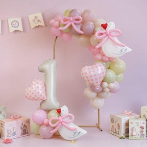 以下が含まれることがあります： ピンクとゴールドをテーマにした、お祝いの1歳の誕生日セット。 バルーンアーチ、大きな数字1のバルーン、ハート型のバルーン、装飾されたギフトボックスが特徴です。 イラスト入りのバナーと白いガチョウのバルーンも含まれています。